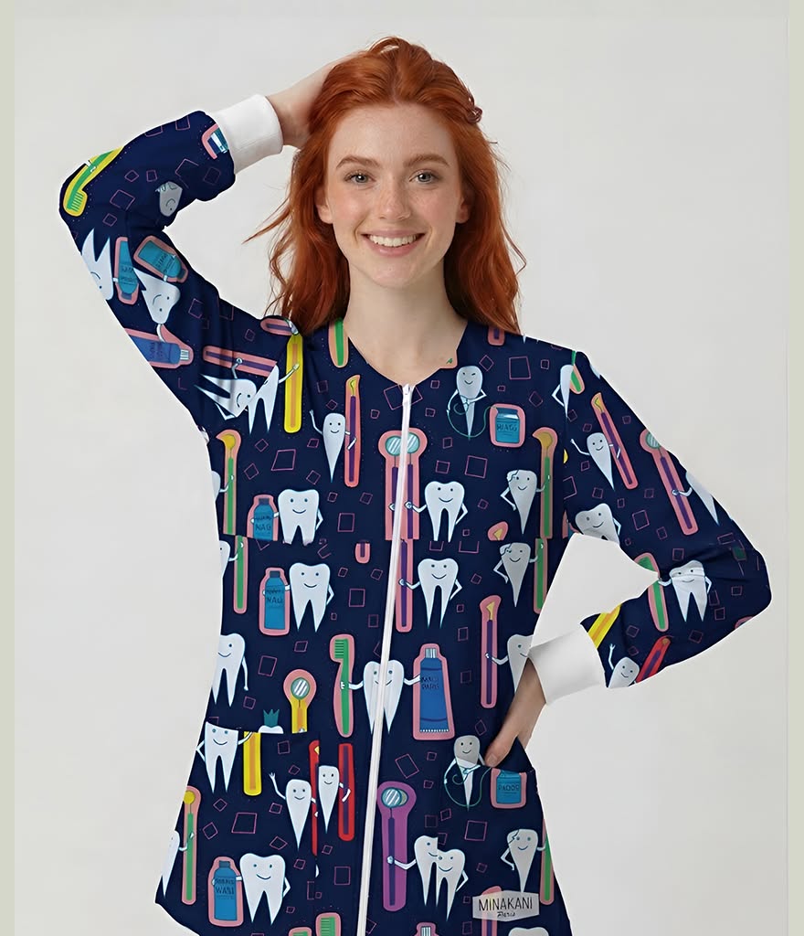 A smiling young woman with red hair wears a navy long-sleeve zip-front scrub top covered in a colorful cartoon tooth and dental tool pattern, photographed against a light gray studio background.
