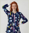 A smiling young woman with red hair wears a navy long-sleeve zip-front scrub top covered in a colorful cartoon tooth and dental tool pattern, photographed against a light gray studio background.