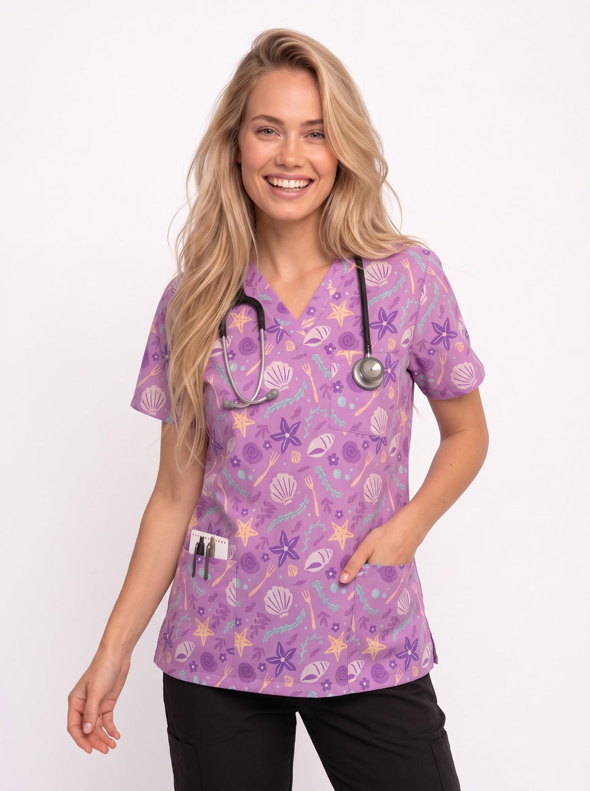A smiling blonde woman wearing a purple short-sleeve scrub top with a seashell and starfish pattern, a black stethoscope around her neck, and black pants, photographed against a clean white studio background.