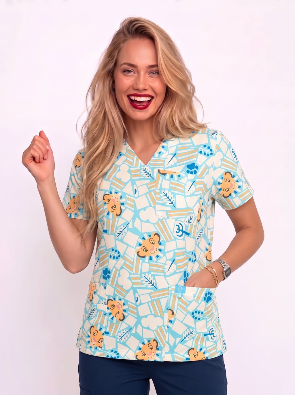 A smiling blonde woman wearing a light blue and white scrub top with a playful cartoon pattern of faces, animals, and medical icons, paired with navy pants, set against a plain white studio background.