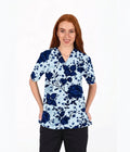 A woman with red hair wearing a light blue and navy blue floral print scrub top, paired with black pants, standing against a plain white background.